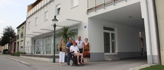 Fünf Personen, darunter eine im Rollstuhl, stehen an einem sonnigen Tag vor einem modernen Gebäude mit Glasfenstern und Balkonen.