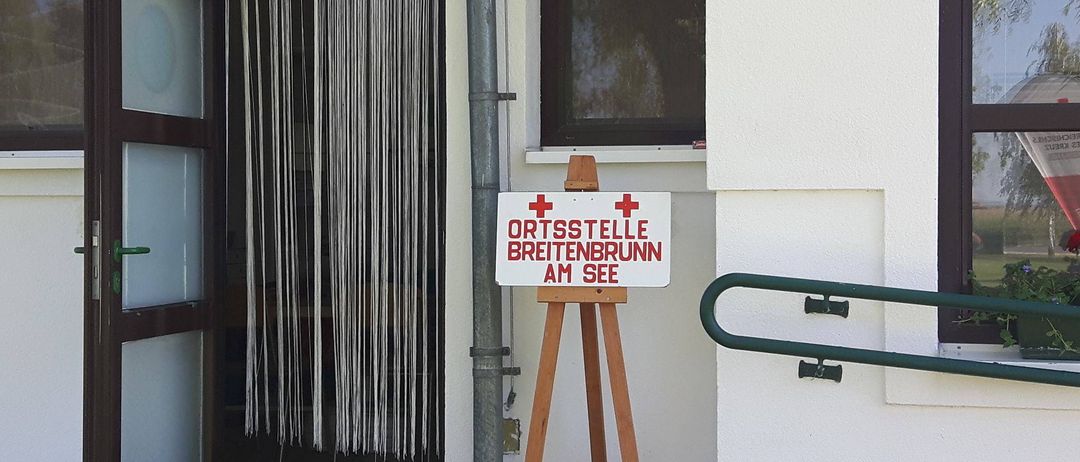 Ein weißes Schild mit roten Kreuzen steht auf einem hölzernen Staffelei vor einem Gebäude. Das Schild zeigt 'ORTSSTELLE BREITENBRUNN AM SEE'. Das Gebäude hat eine weiße Wand, ein Fenster mit schwarzem Rahmen und eine Metallrohre auf der linken Seite. Das Fenster ist leicht geöffnet.