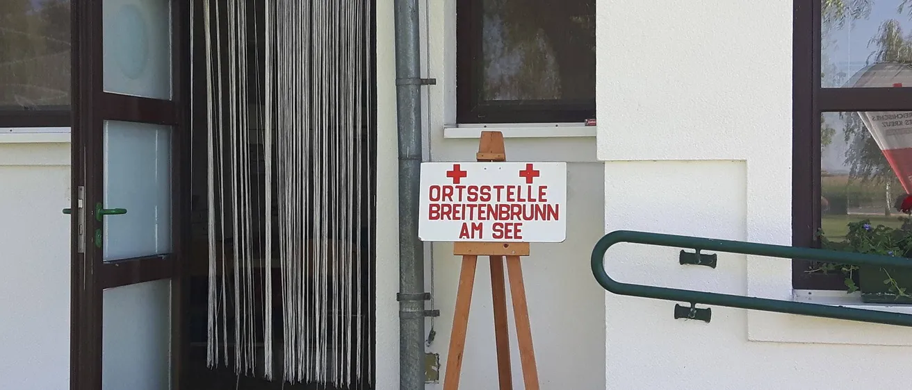 Ein weißes Schild mit roten Kreuzen steht auf einem hölzernen Staffelei vor einem Gebäude. Das Schild zeigt 'ORTSSTELLE BREITENBRUNN AM SEE'. Das Gebäude hat eine weiße Wand, ein Fenster mit schwarzem Rahmen und eine Metallrohre auf der linken Seite. Das Fenster ist leicht geöffnet.