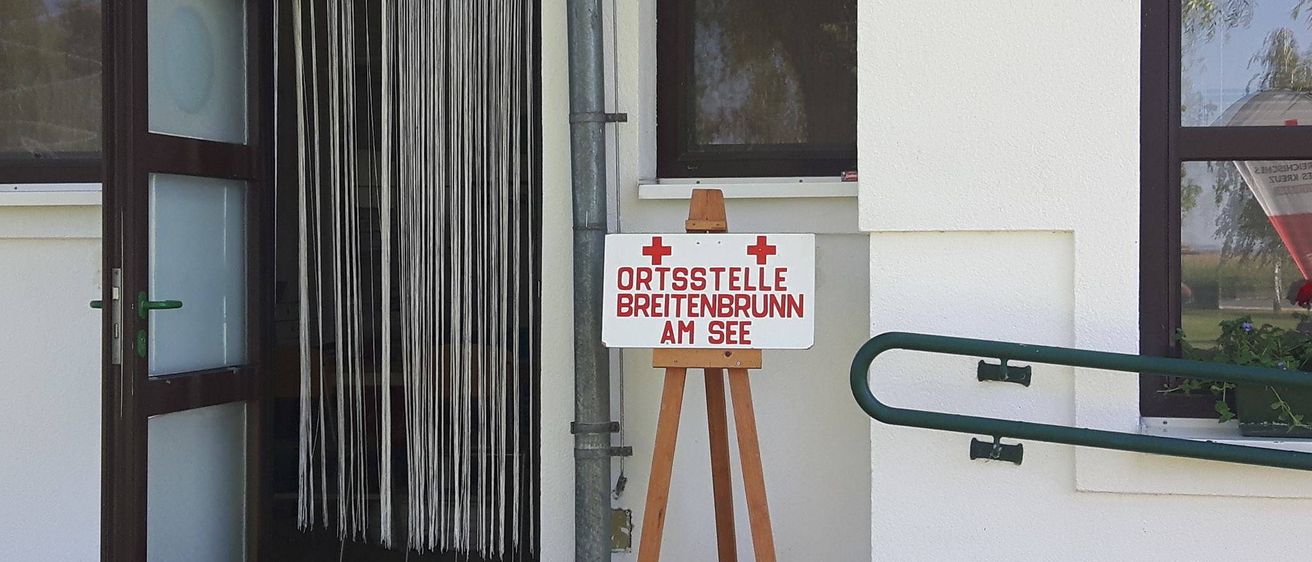 Ein weißes Schild mit roten Kreuzen steht auf einem hölzernen Staffelei vor einem Gebäude. Das Schild zeigt 'ORTSSTELLE BREITENBRUNN AM SEE'. Das Gebäude hat eine weiße Wand, ein Fenster mit schwarzem Rahmen und eine Metallrohre auf der linken Seite. Das Fenster ist leicht geöffnet.