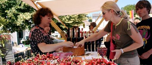 Eine Frau kauft Kirschen von einem Verkäufer unter einem Vordach auf einem Outdoor-Markt. Mehrere Behälter mit Kirschen sind ausgestellt, ein Flagge ist im Hintergrund sichtbar.