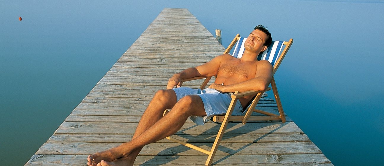 Ein Mann ruht sich auf einer Holzplattform in der Nähe eines Sees aus, trägt Shorts und einen Sonnenstuhl. Der Mann liegt mit geschlossenen Augen da.