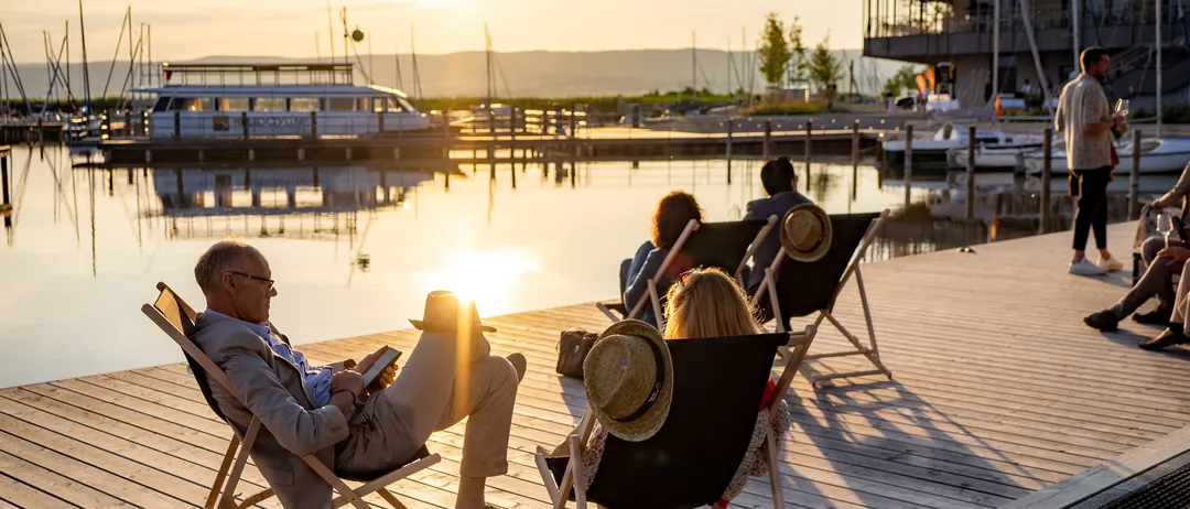 Mehrere Menschen entspannen sich auf einer Holzterrasse in der Nähe eines Sees, mit Booten und einem Gebäude im Hintergrund. Die Sonne geht unter.