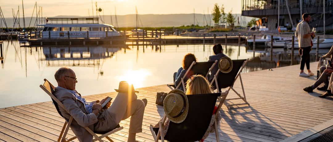 Mehrere Menschen entspannen sich auf einer Holzterrasse in der Nähe eines Sees, mit Booten und einem Gebäude im Hintergrund. Die Sonne geht unter.