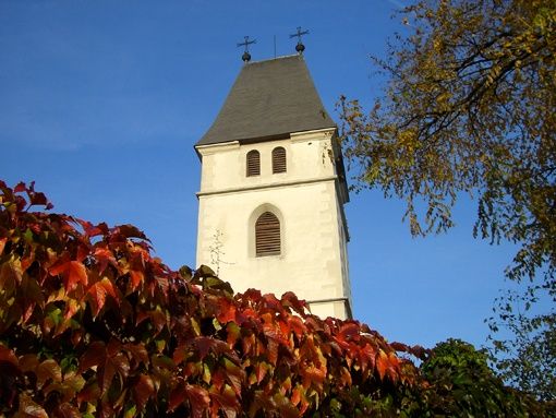 Ein weißer Kirchturm mit zwei Kreuzen auf der Spitze, umgeben von Bäumen mit roten Blättern gegen einen blauen Himmel.