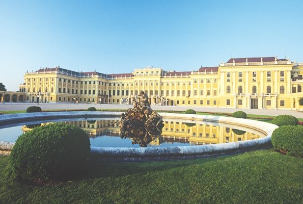 Ein großes gelbes Schloss mit vielen Fenstern und einem Brunnen davor an einem sonnigen Tag.