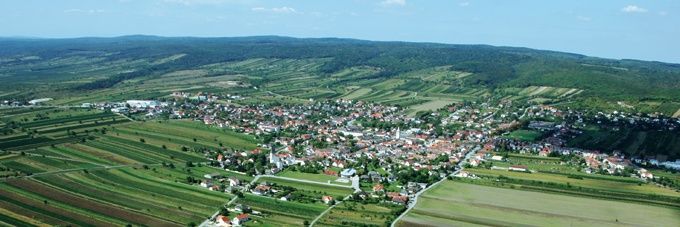 Luftaufnahme eines Dorfes umgeben von grünen Feldern und Hügeln unter einem blauen Himmel mit Wolken.