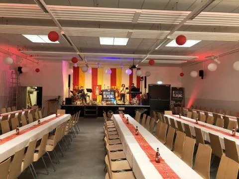 Ein großer Raum mit Bühne und Stühlen für eine Veranstaltung. Die Bühne hat einen rot-gelben Hintergrund mit Luftballons. Die Tische sind mit weißen Tischdecken und roten Tischläufern bedeckt. Flaschen stehen auf den Tischen.