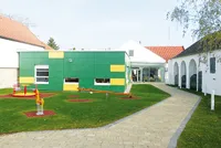 Ein leuchtend grünes und gelbes Gebäude steht inmitten eines grünen Bereichs mit Spielplatz, gepflastertem Weg und roten Dächern.