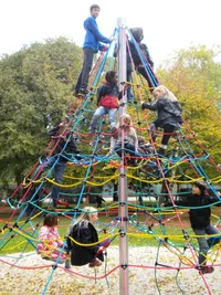 Mehrere Kinder klettern in einem Outdoor-Park an einer farbenfrohen Netzstruktur. Bäume sind im Hintergrund zu sehen.