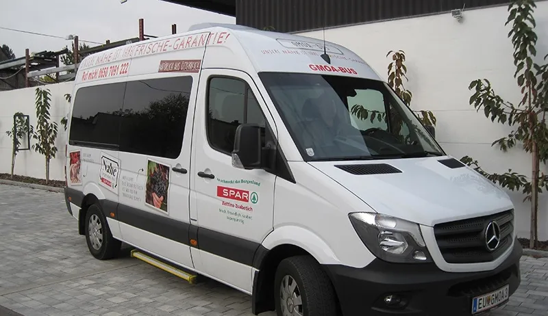 Ein weißer Mercedes-Benz-Van mit Werbung an der Seite steht auf einer gepflasterten Fläche in der Nähe eines Gebäudes. Er hat das Kennzeichen 'EU-GMOA' und den Namen 'Gmoa-Bus' an der Seite.