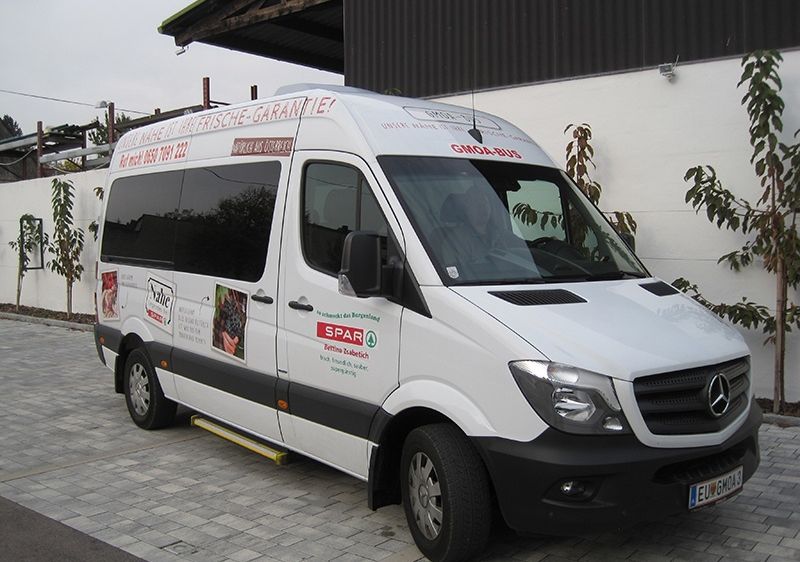 Ein weißer Mercedes-Benz-Van mit Werbung an der Seite steht auf einer gepflasterten Fläche in der Nähe eines Gebäudes. Er hat das Kennzeichen 'EU-GMOA' und den Namen 'Gmoa-Bus' an der Seite.