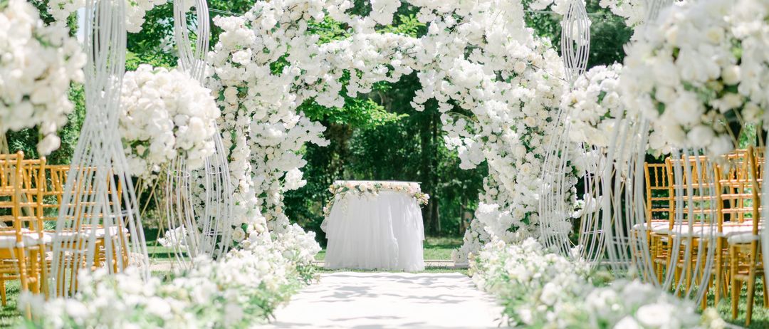 Ein Hochzeitsgang, der mit weißen Blumen und Grün bedeckt ist, mit einem weißen Tisch in der Mitte. Im Hintergrund sind Bäume und Grün zu sehen.