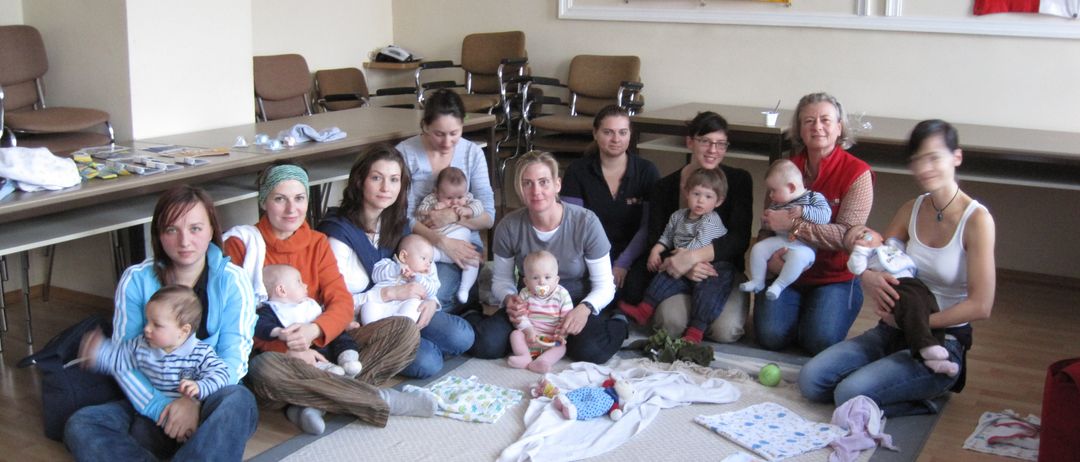 Eine Gruppe von Frauen und Kindern versammelt sich in einem Raum. Sie sitzen auf dem Boden und halten Babys. Es gibt Babykleidung auf dem Boden und eine Flagge an der Wand.