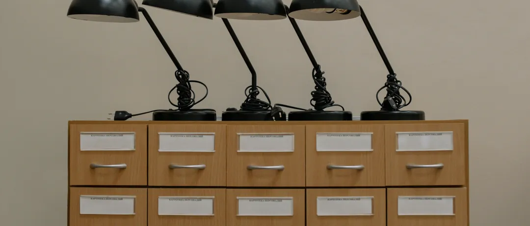 Vier schwarze Schreibtischlampen sind auf einem Holzschrank angeordnet. Jede Lampe hat einen schwarzen Sockel und einen schwarzen Arm. Die Lampen sind an eine Steckdose angeschlossen.