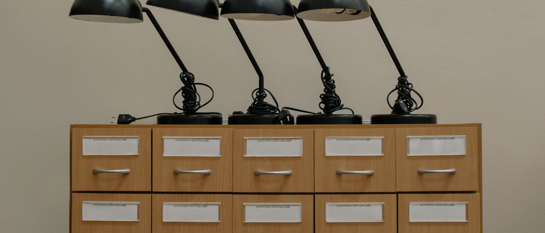 Vier schwarze Schreibtischlampen sind auf einem Holzschrank angeordnet. Jede Lampe hat einen schwarzen Sockel und einen schwarzen Arm. Die Lampen sind an eine Steckdose angeschlossen.