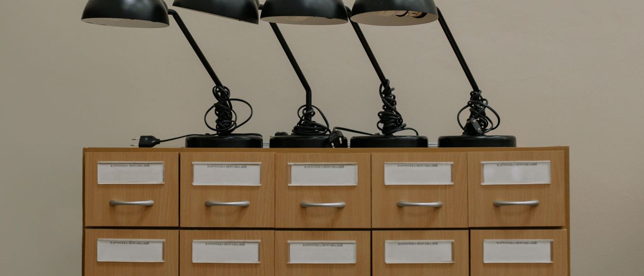 Vier schwarze Schreibtischlampen sind auf einem Holzschrank angeordnet. Jede Lampe hat einen schwarzen Sockel und einen schwarzen Arm. Die Lampen sind an eine Steckdose angeschlossen.