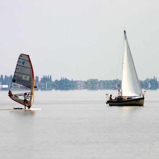 Zwei Segelboote auf einer ruhigen Wasserfläche, eines mit einem Windsurfer und das andere mit einer sitzenden Person.
