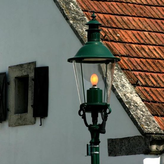 Eine grüne Straßenlaterne mit einer leuchtenden gelben Glühbirne ist an der Wand eines weißen Gebäudes montiert. Das Dach besteht aus roten Ziegeln. Neben der Laterne befindet sich ein Fenster mit zwei Fensterläden.