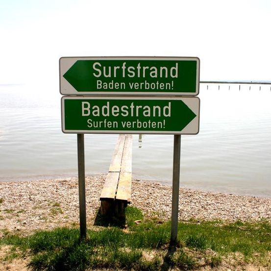 Zwei Schilder am Strand. Das obere Schild sagt Surfstrand und zeigt an, dass Schwimmen verboten ist. Das untere Schild sagt Badestrand und zeigt ebenfalls an, dass Surfen verboten ist.