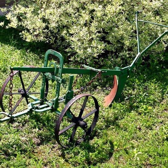 Ein alter grüner Pflug mit zwei Rädern und einem Metallgriff steht auf dem Gras. Er ist mit einer Kette verbunden.
