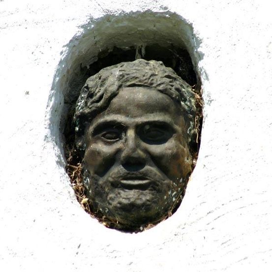 Eine verwitterte Steinskulptur eines Männerkopfes ragt aus einer weißen Wand hervor, mit sichtbaren Rissen und Schmutz.