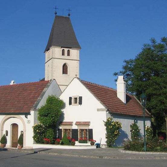 Eine weiße Kirche mit einem braunen Dach und einem Turm, umgeben von einem Garten mit gelben Blumen und grünen Pflanzen. Es gibt zwei Topfpflanzen an der Seite des Eingangs.