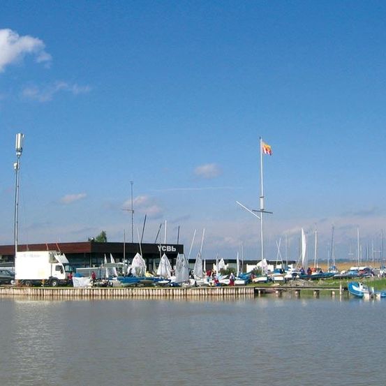 Ein Außenbereich mit einem großen Gebäude hat viele Segelboote, die am Wasser verankert sind. Ein Lkw ist in der Nähe geparkt, und eine Flagge ist auf einem Mast gehisst. Einige Leute stehen auf der Dock.