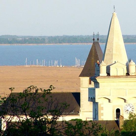 Ein hoher Kirchturm mit einer Uhr ist in der Ferne zu sehen, umgeben von einem weiten, offenen Feld und einem ruhigen See mit angedockten Booten.