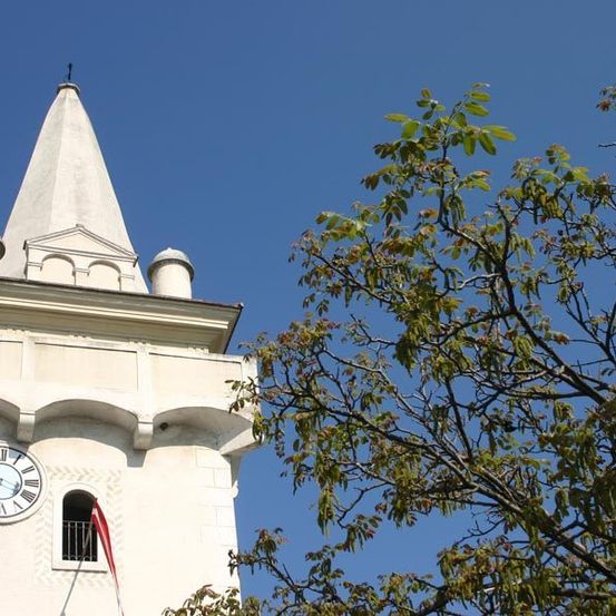 Ein hoher weißer Glockenturm mit einem Spitzturm, der eine Uhr und eine Fahne unter einem Fenster zeigt, steht gegen einen klaren blauen Himmel mit einem Baum im Vordergrund.