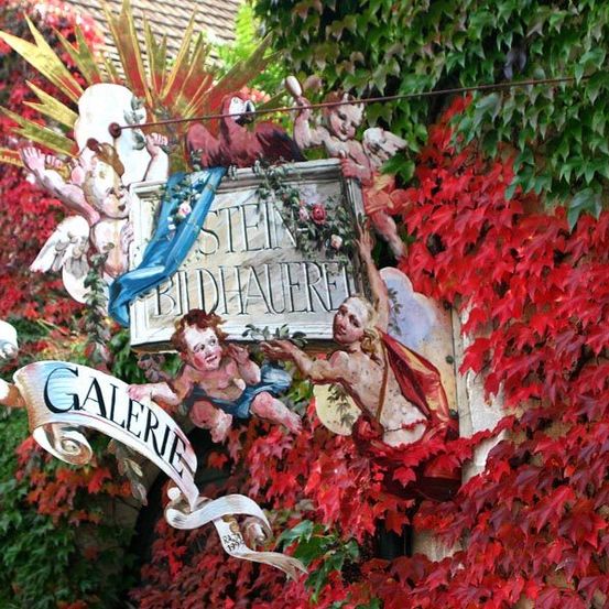 Ein Schild für Galerie Stein Bidhauerei ist an einer mit roten und grünen Blättern bedeckten Wand montiert. Es zeigt Cherubinen und einen Papagei.
