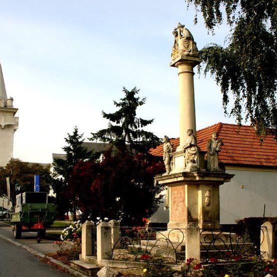 Ein Denkmal mit Statuen steht vor einer Kirche, mit einem Glockenturm und einem grünen Wagen auf der Straße.
