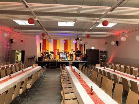 Ein großer Innenbereich mit Bühne, roten und gelben Vorhängen und für eine Party gedeckten Tischen. Flaschen und Luftballons stehen auf den Tischen.