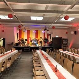 Ein großer Innenbereich mit Bühne, roten und gelben Vorhängen und für eine Party gedeckten Tischen. Flaschen und Luftballons stehen auf den Tischen.