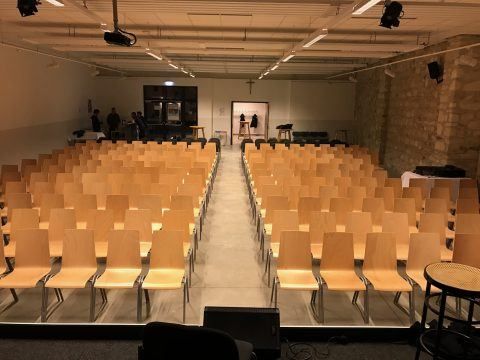 Ein leerer Konferenzraum mit Reihen von Holzstühlen und einem Mittelgang. Der Raum hat eine Bühne, Tische und Beleuchtungsgeräte. Im Hintergrund befinden sich Personen und ein Kreuz ist an der Wand sichtbar.
