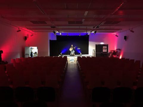 Ein leerer Saal ist für eine Aufführung vorbereitet. Eine Person steht mit einer Gitarre auf der Bühne, wobei rote und blaue Lichter den Bereich beleuchten.
