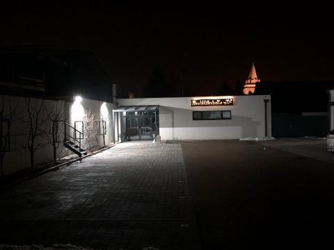Ein Gebäude ist nachts beleuchtet. Der Eingang hat ein Vordach und Treppen. Ein gelbes Schild mit 'Schillerhof' ist zu sehen. Ein Turm ist in der Ferne sichtbar.