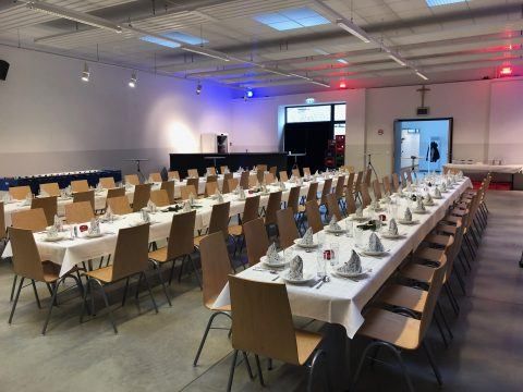 Ein großer Bankettsaal mit mehreren langen Tischen, die für ein Dinner-Event vorbereitet sind. Jeder Tisch hat Teller, Servietten und Besteck. Der Raum hat weiße Wände, Deckenleuchten und blaue Lichter an den Wänden.