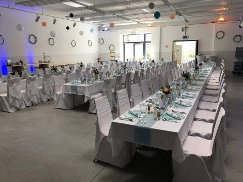 Ein Bankettsaal ist für eine Veranstaltung vorbereitet mit langen Tischen, die mit weißen und blauen Tischdecken bedeckt sind, jeweils mit Blumen, Tellern und Besteck. Weiße Stühle sind um die Tische herum angeordnet. Der Raum ist mit Luftballons und Kränzen an der Wand dekoriert.