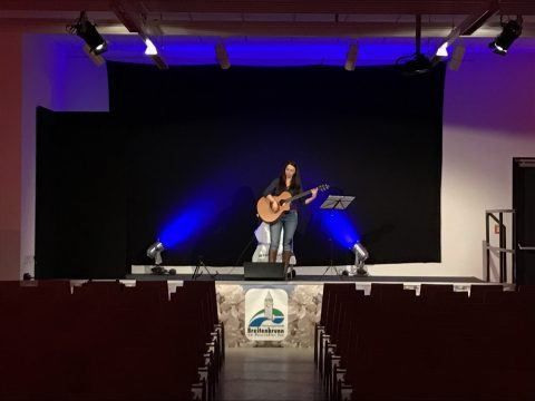 Eine Frau steht mit einer Gitarre und einem Mikrofon auf der Bühne, sie spielt vor einer schwarzen Kulisse und blauen Beleuchtung. Die Bühne ist leer, mit Reihen von Stühlen, die auf sie gerichtet sind.