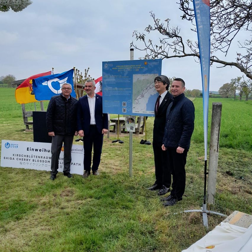 Vier Männer stehen vor einem Schild mit Flaggen und einer Karte. Zwei Banner stehen hinter ihnen, eines mit einer Kirschblüte und das andere mit dem Text 'Kirschblütenradweg'.