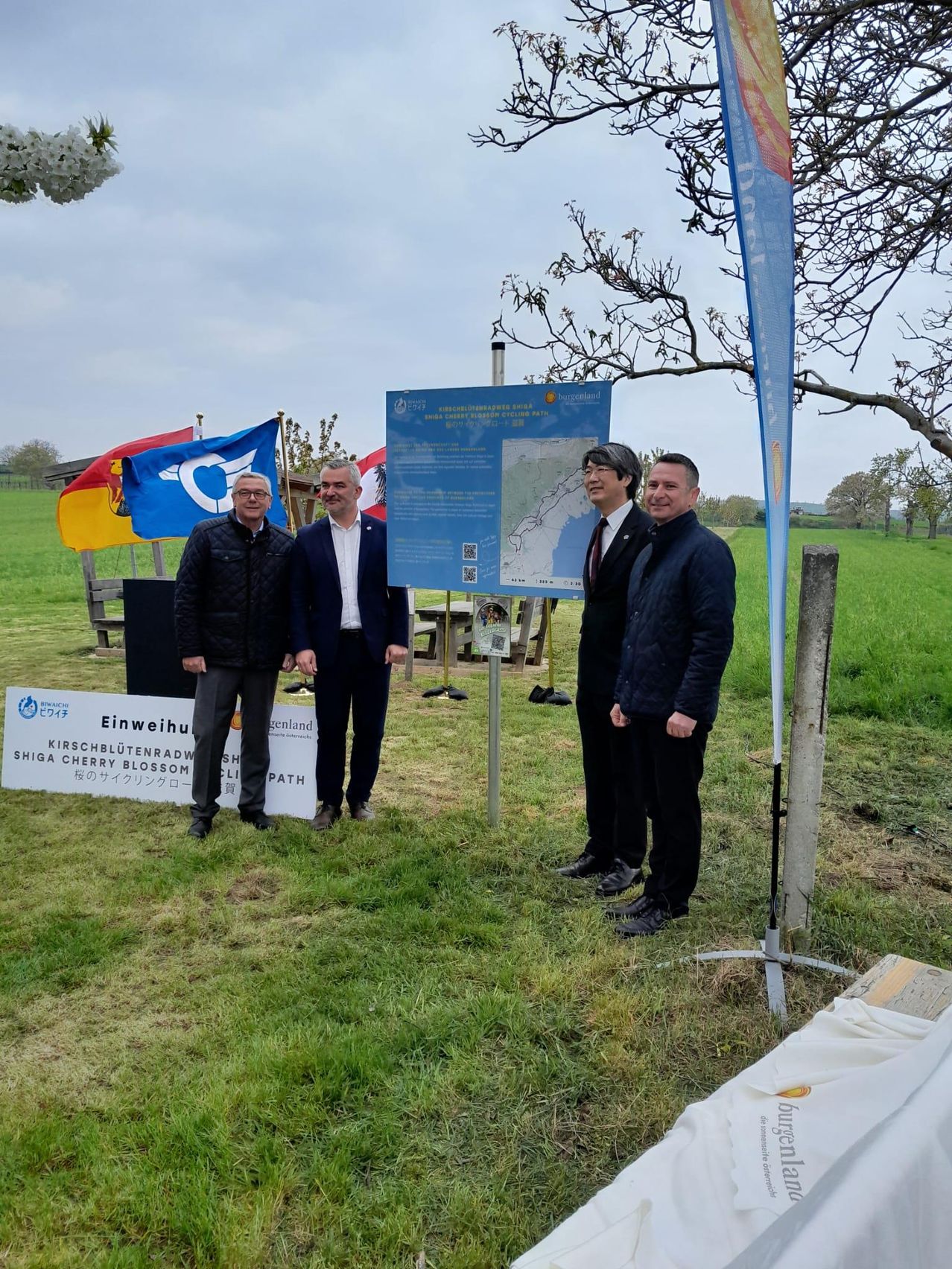 Vier Männer stehen vor einem Schild mit Flaggen und einer Karte. Zwei Banner stehen hinter ihnen, eines mit einer Kirschblüte und das andere mit dem Text 'Kirschblütenradweg'.