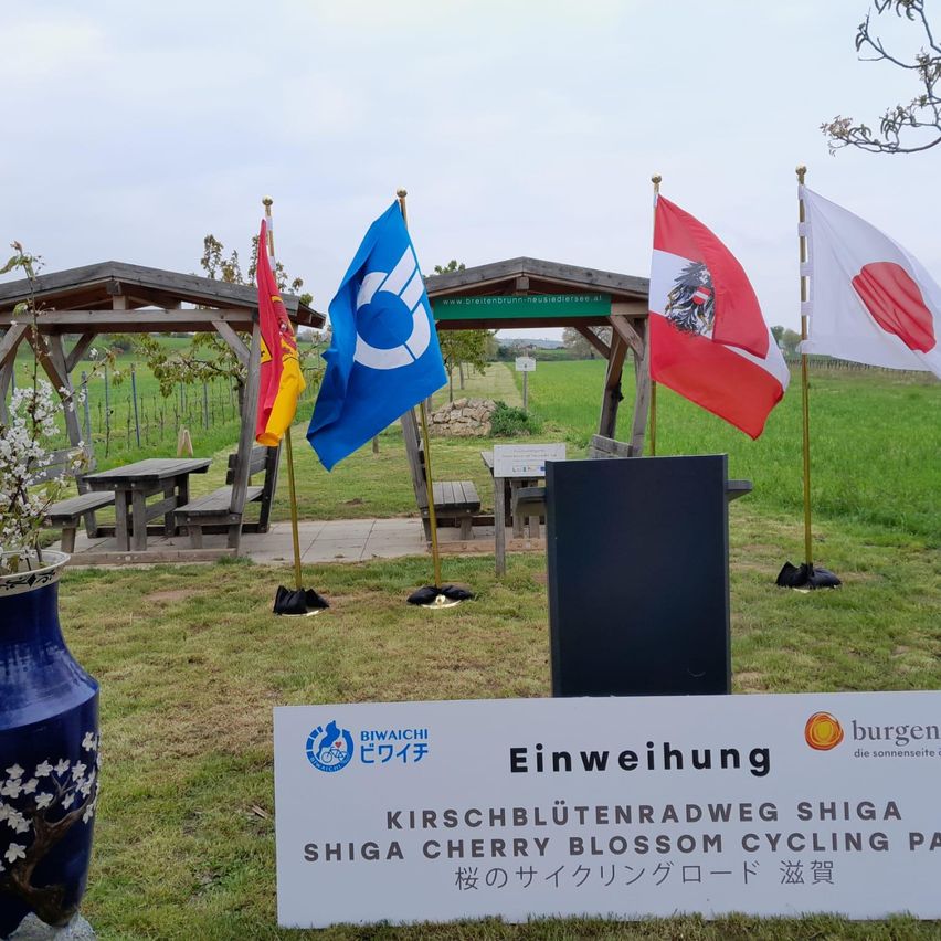 Eine Zeremonie am Eingang des Shiga Kirschblüten-Radwegs mit Flaggen verschiedener Länder und einer blauen Vase mit weißen Blumen.