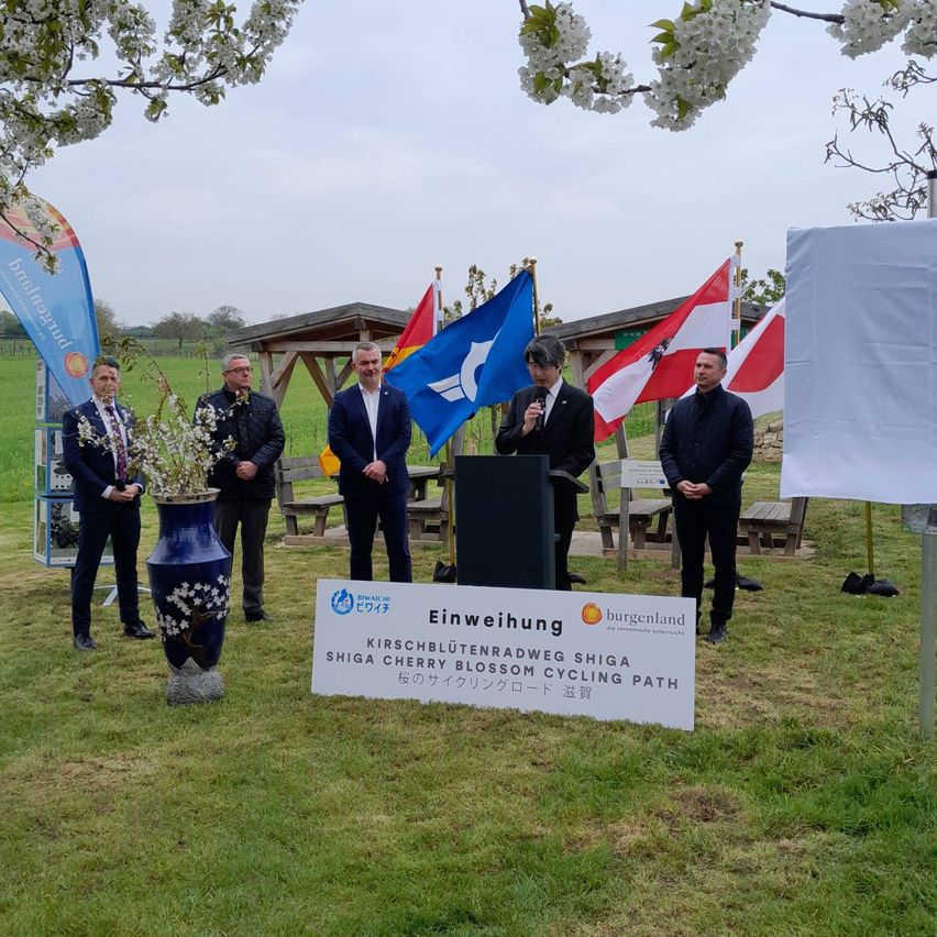 Offizielle stehen an einem Podium mit Flaggen dahinter und einem Schild für den Shiga Cherry Blossom Cycling Path.