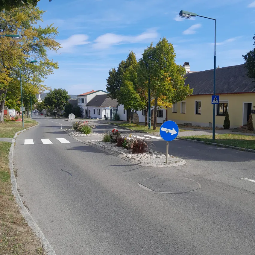 Eine ruhige Straße mit einem blauen Schild, das eine Rechtskurve anzeigt, umgeben von Bäumen und Gebäuden. Die Straße ist leer, und der Himmel ist klar.