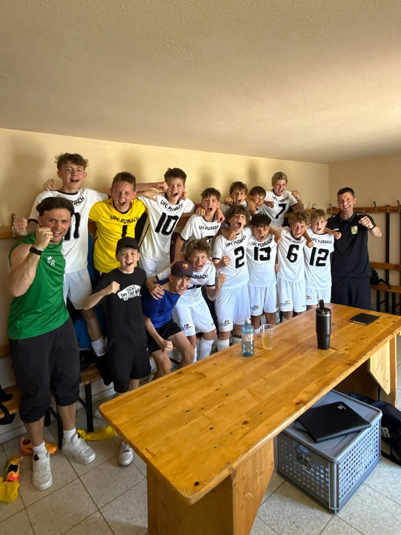 Eine Fußballmannschaft posiert für ein Foto in einer Umkleidekabine, trägt weiße Trikots mit den Nummern 2, 6, 10, 12, 13 und 18. Ein Mann in einem grünen Shirt steht links, und ein anderer in Schwarz steht rechts. Ein Holztisch mit einer Flasche und einem Glas steht davor.