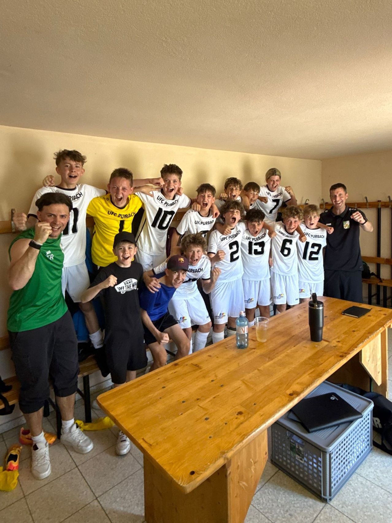 Eine Fußballmannschaft posiert für ein Foto in einer Umkleidekabine, trägt weiße Trikots mit den Nummern 2, 6, 10, 12, 13 und 18. Ein Mann in einem grünen Shirt steht links, und ein anderer in Schwarz steht rechts. Ein Holztisch mit einer Flasche und einem Glas steht davor.