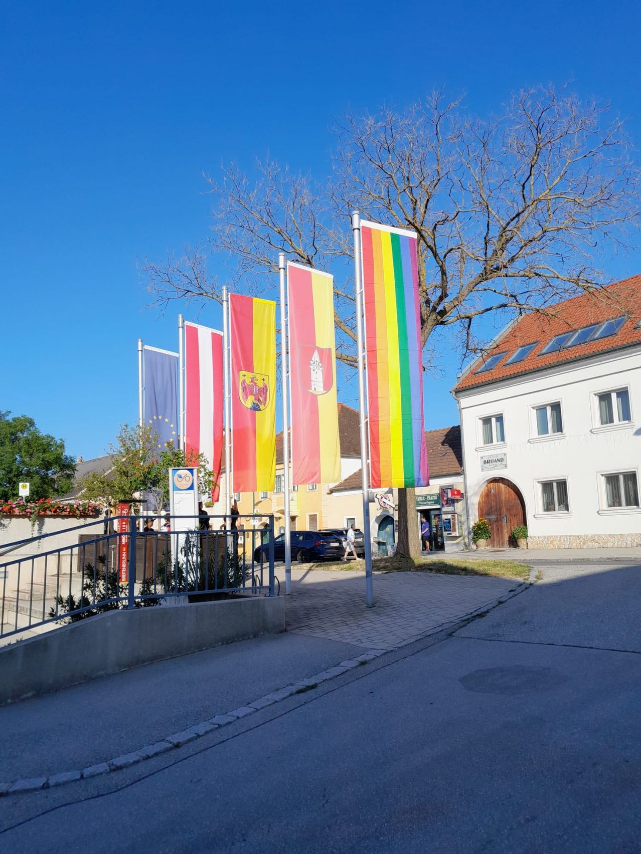 Eine farbenfrohe Straße mit bunten Fahnen und Gebäuden. Bäume und ein Auto sind im Hintergrund zu sehen.