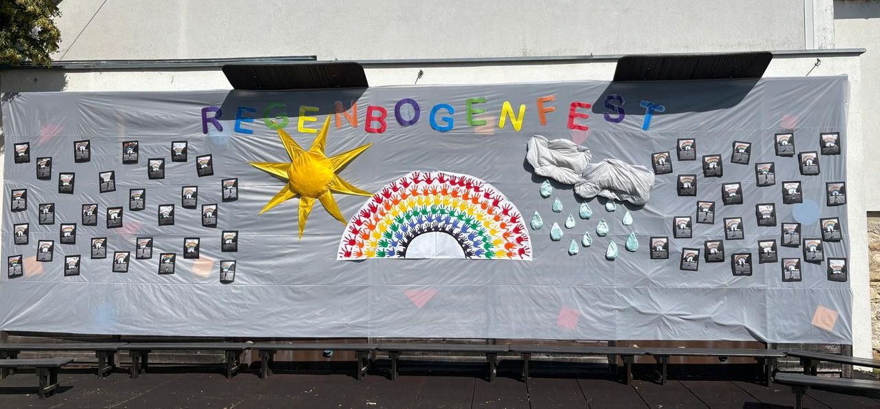 A colorful banner reads 'Egenbogenfest' in various colors. It features a large rainbow, a sun, and some raindrops. The banner is likely for an event.