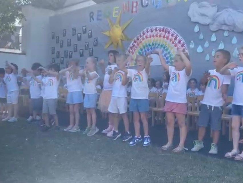 Kinder in weißen T-Shirts mit Regenbogen-Designs stehen in einer Reihe im Freien, Hände erhoben, vor einer Wand mit einem Regenbogen und Wettersymbolen.